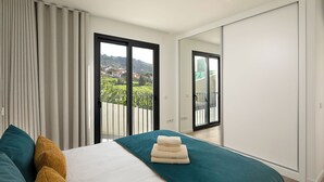 Quarto com cama, cortinas, vista por uma porta deslizante e varanda.