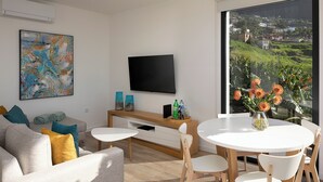 Uma sala de estar moderna com televisão de tela plana, sofá, mesa de jantar com cadeiras e uma grande janela com vista para uma paisagem verde.