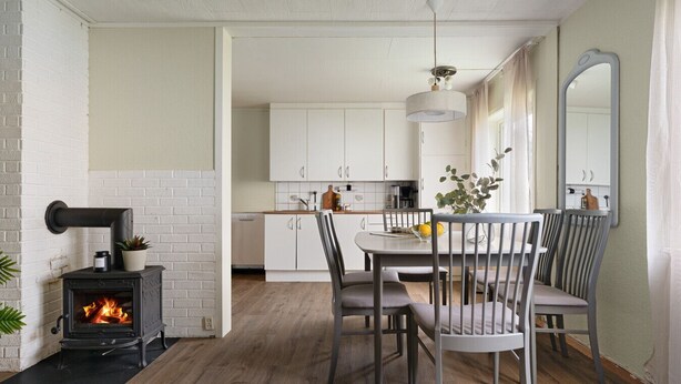 Et moderne køkken med hvide skabe, et spisebord med stole, en sort brændeovn og en hængende lampe.