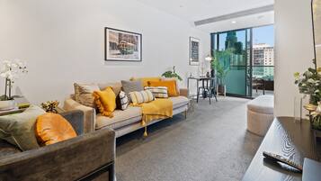 A modern living room with a sofa, ottoman, and coffee table, a dining area with a table and chairs, and large windows offering a city view.