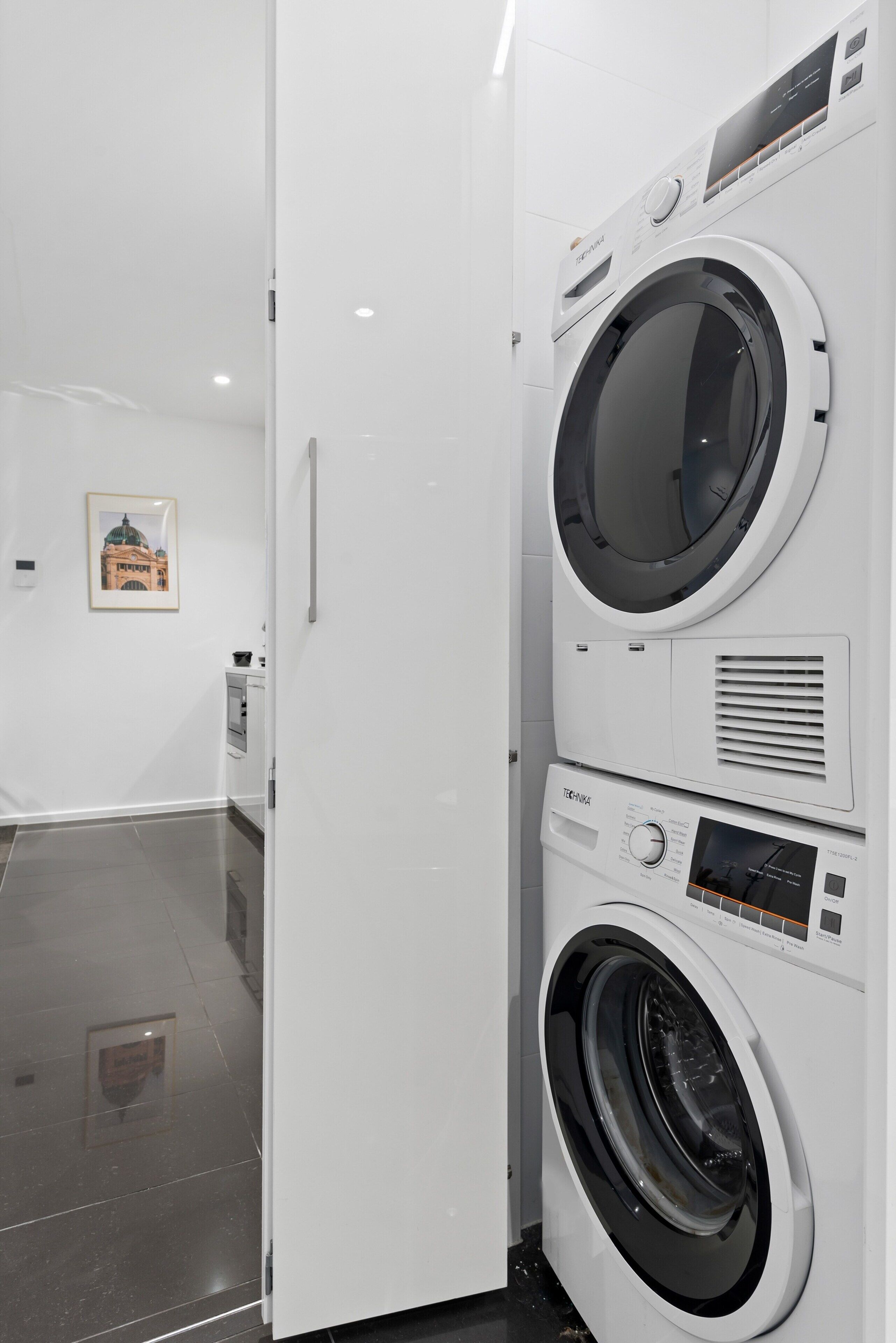A modern laundry room with two stacked front-loading washing machines, a digital display, and a small kitchenette area with a stove and microwave.