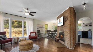 A spacious living room with hardwood flooring, a fireplace, a ceiling fan, and a large window with curtains. - CozyCottage_Sleeps8_Near Plaza and KU_and Shopping (Mission)