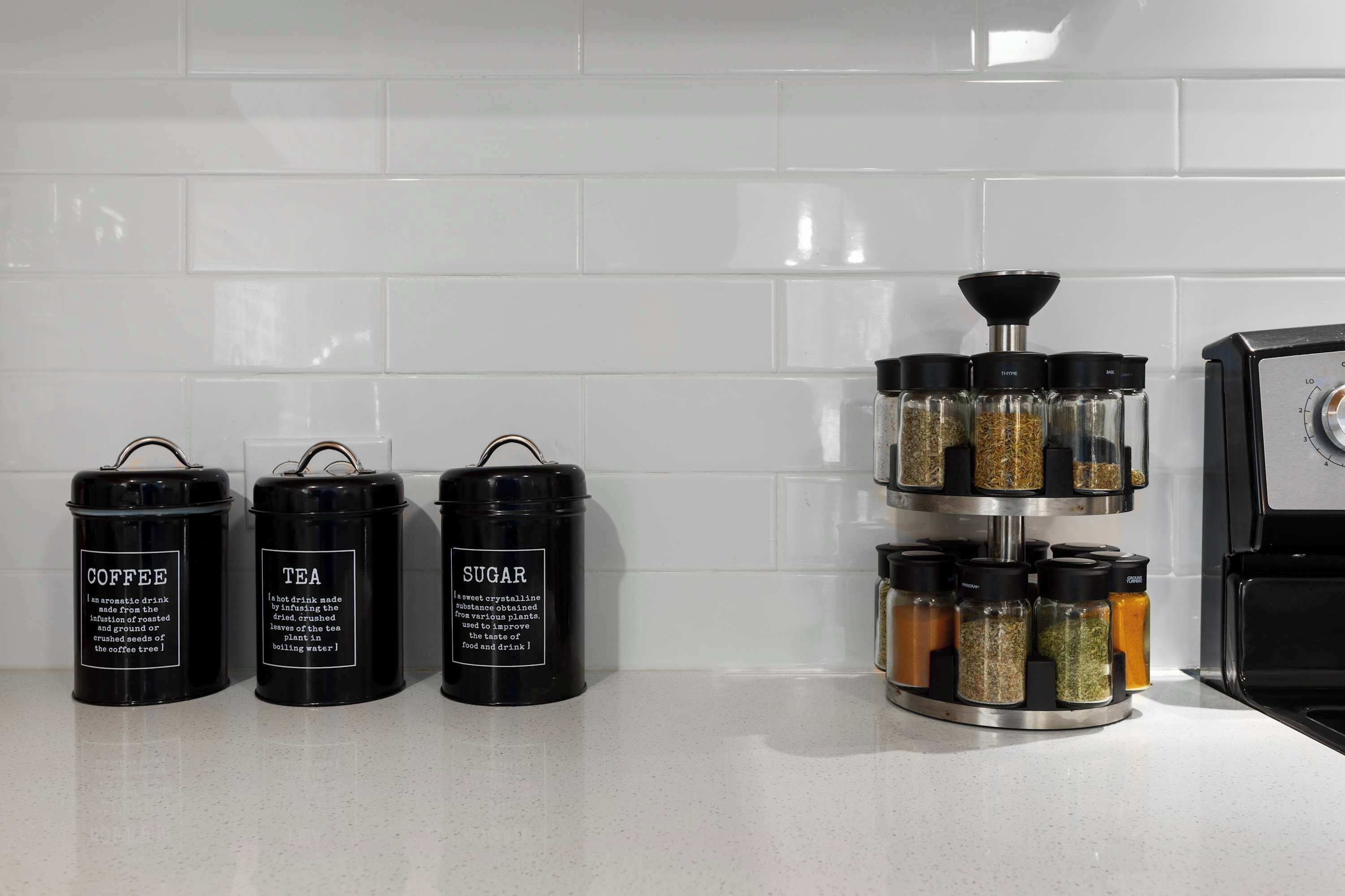 Tre contenitori neri etichettati caffè, tè e zucchero su un piano di lavoro della cucina con zanzariera bianca.