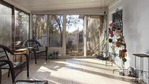 A sunlit patio with wicker chairs, a glass door leading outside, and a metal shelf with potted plants. - CozyCottage_Sleeps8_Near Plaza and KU_and Shopping (Mission)
