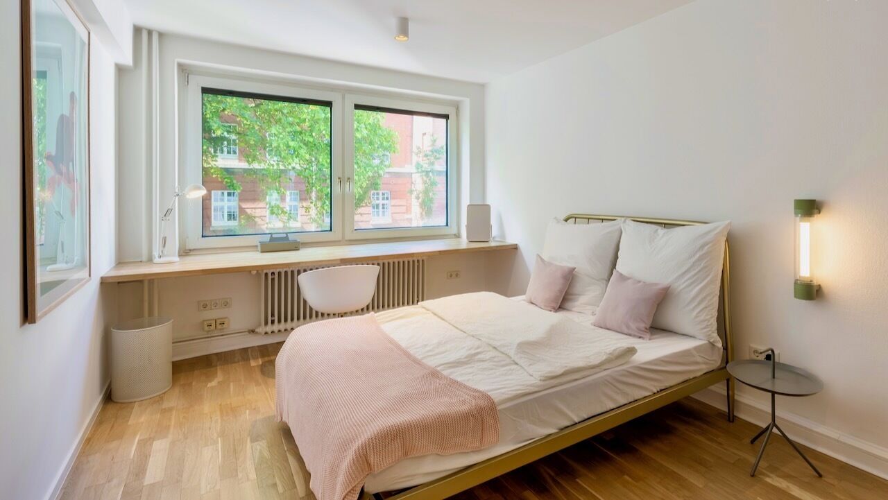 A bedroom with a bed, a desk, a radiator, and a window with a view of trees and buildings.