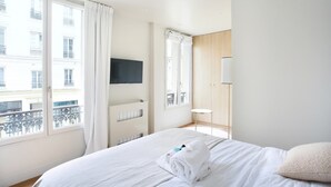 Habitación de hotel con cama, televisor montado en la pared, ventana con cortinas y vistas a edificios.