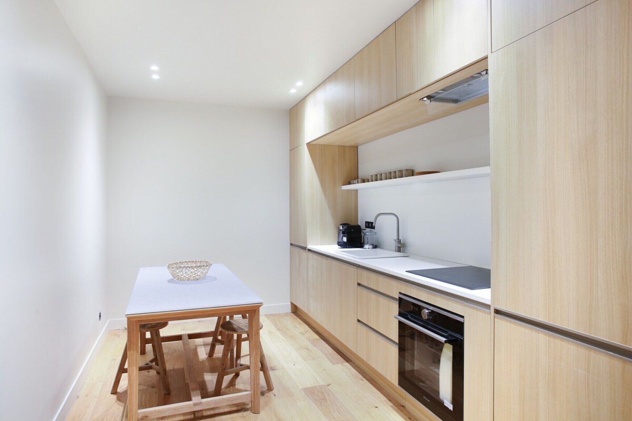 A compact kitchen with a white table, wooden stools, a coffee maker, and a built-in oven.