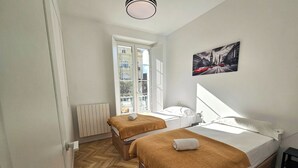 A hotel room with two beds, a radiator, a window with a view of buildings, and a painting on the wall. - Apartamentos Ciudadela Superior 3000 (Jaca)