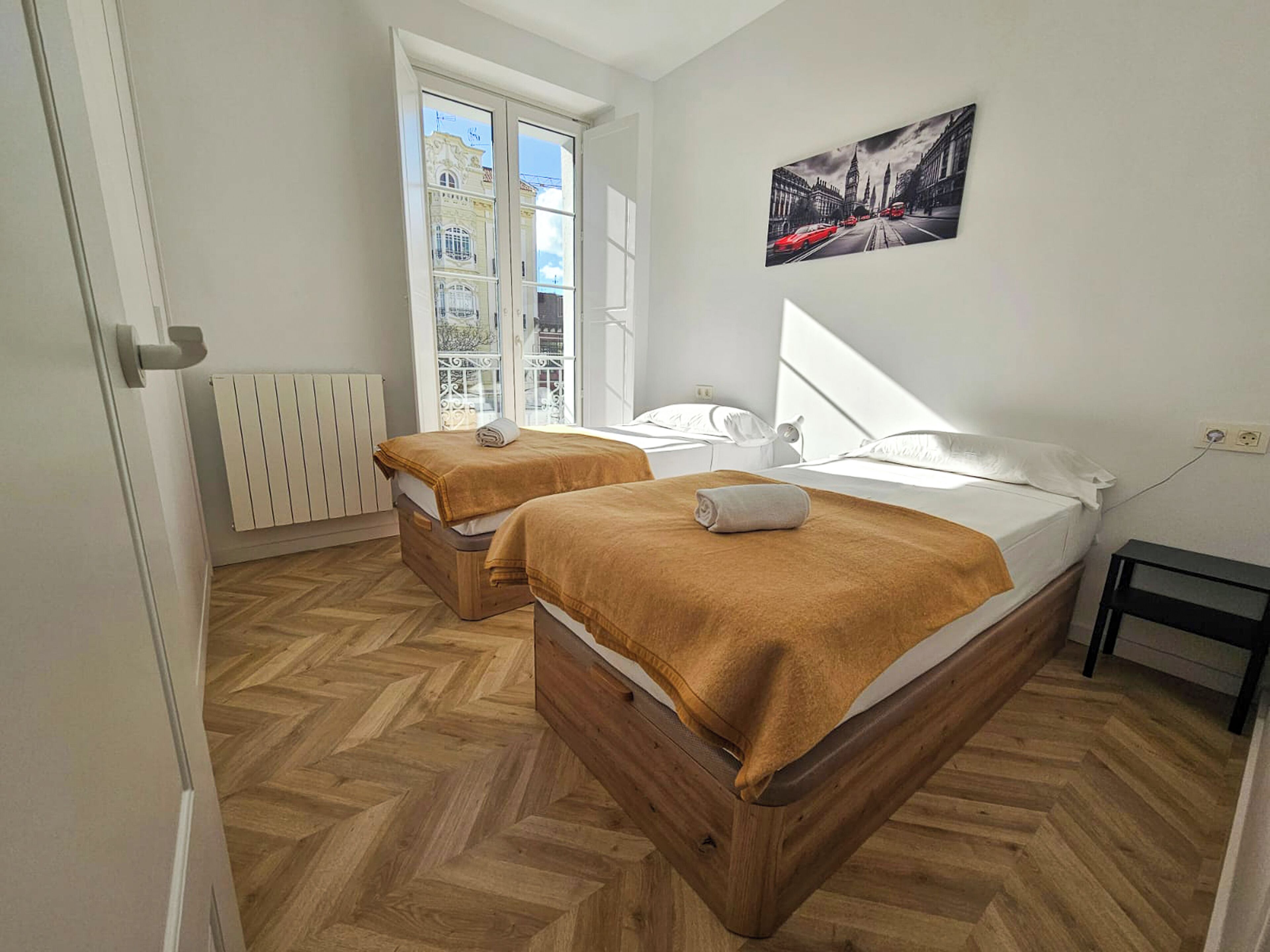 A room with two beds, a radiator, a window with a view of buildings, and a painting on the wall.