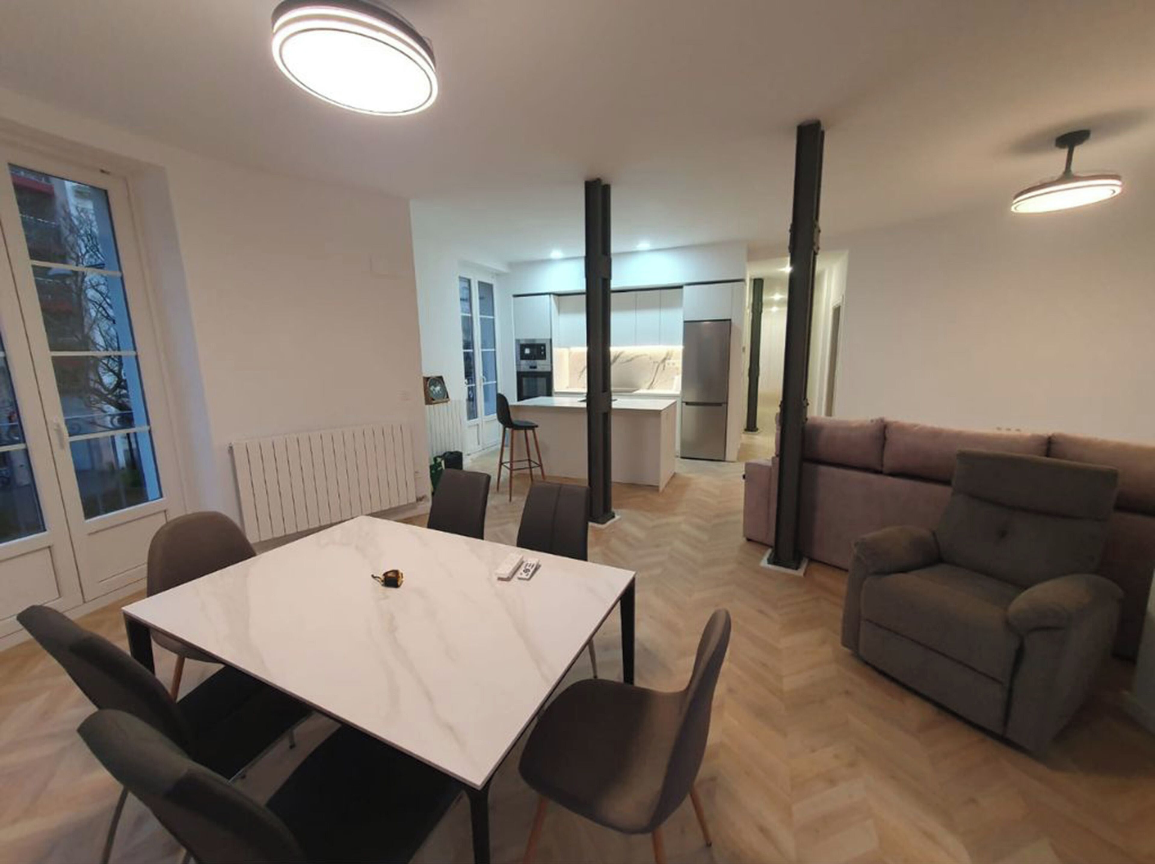 A modern living room with a white marble table, black chairs, a grey sofa, and a kitchen in the background.