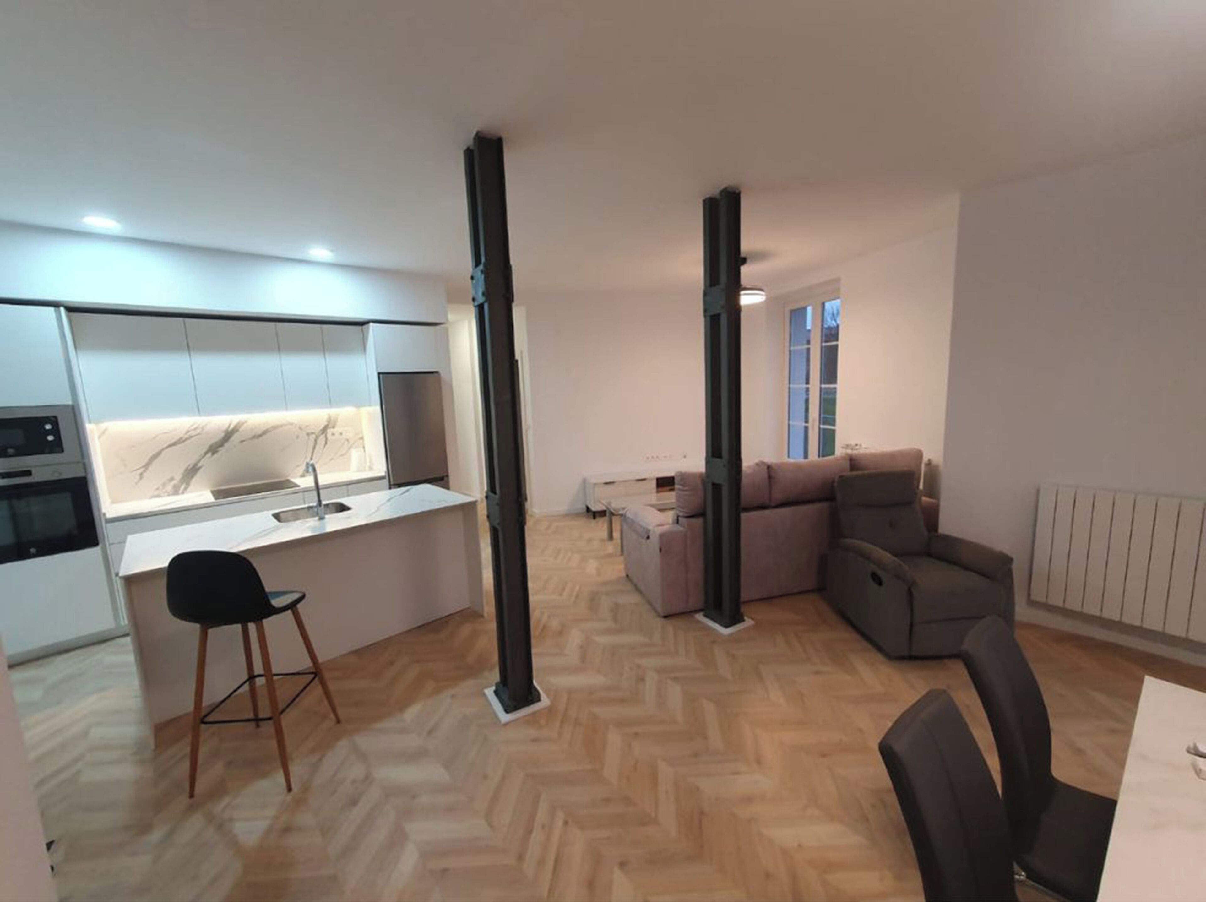 Una cocina moderna con armarios blancos y un salpicadero de mármol, una zona de comedor con mesa y sillas, y un salón con sofá y radiador.