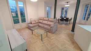 A spacious living area with a large L-shaped sofa, a glass coffee table, and a dining table with chairs. There are large windows providing natural light and views of the outdoors. - Apartamentos Ciudadela Superior 3000 (Jaca)