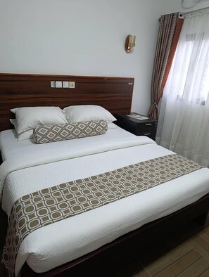 A neatly made bed with a patterned bedspread and pillows, a wooden headboard, a bedside table with a lamp, and a window with curtains. - GUIM'S SQUARE HOTEL (Douala)