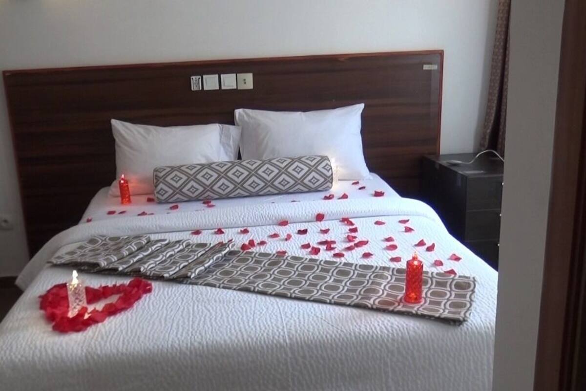 A neatly made bed with white bedding, a patterned pillow, and rose petals scattered across the surface.
