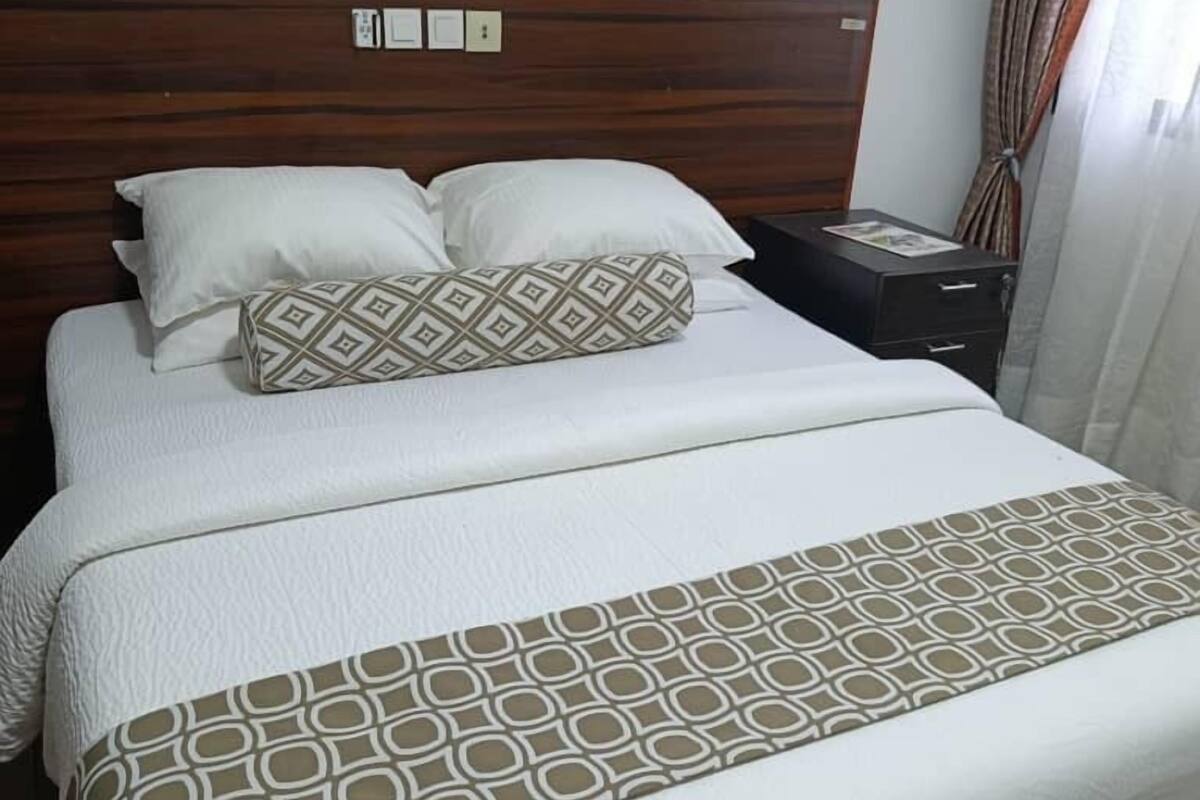 A neatly made bed with a patterned bedspread and pillows, a wooden headboard, a nightstand with a lamp, and a window with curtains.