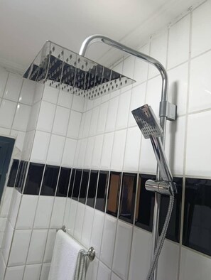 A shower with a transparent rain showerhead and a handheld showerhead, set against a tiled wall with a black horizontal stripe. - GUIM'S SQUARE HOTEL (Douala)