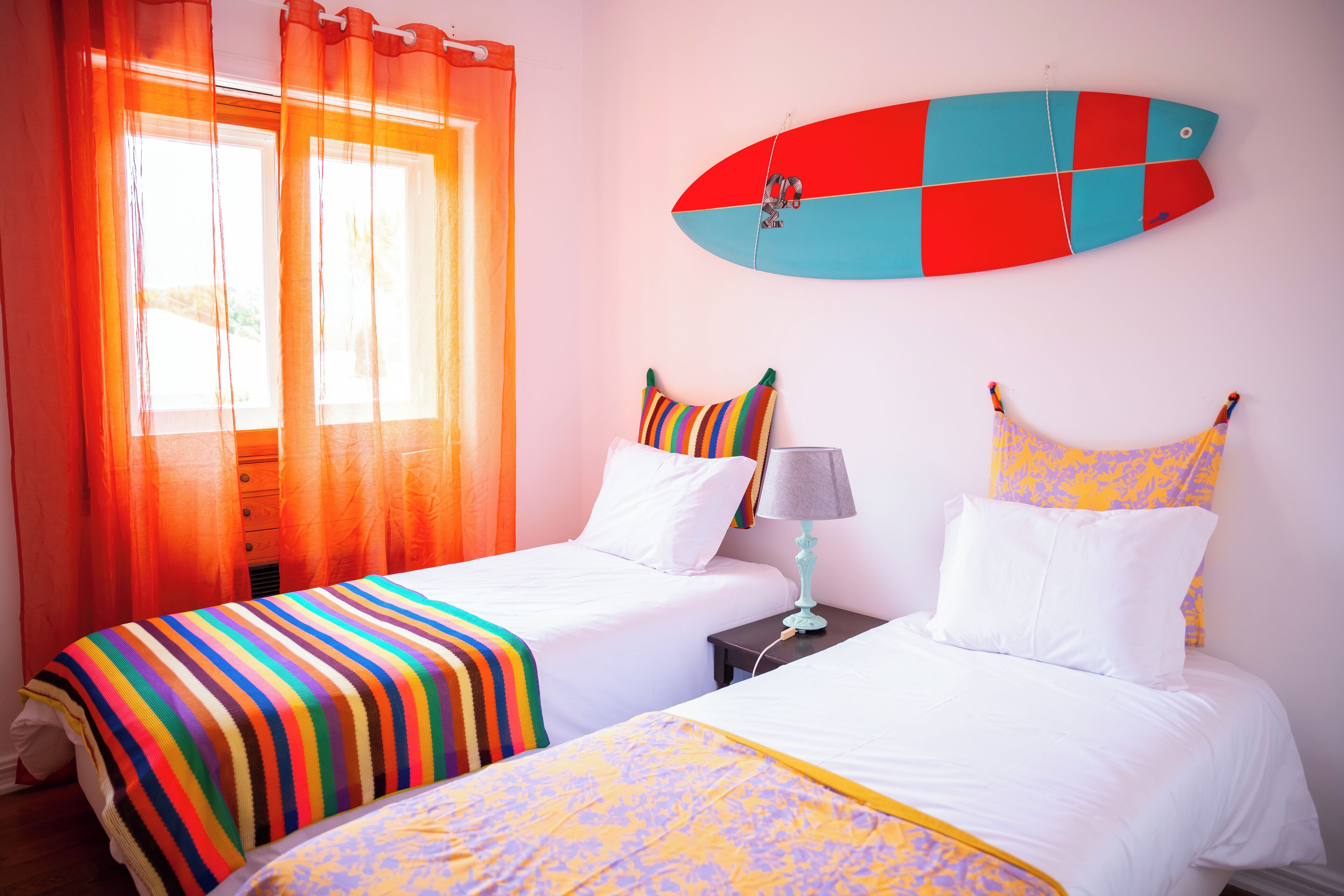 Una habitación con dos camas, una tabla de surf en la pared y una ventana con cortinas naranjas.