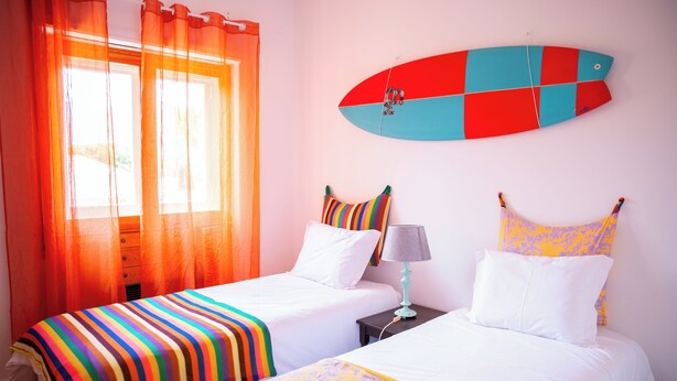 A room with two beds, a surfboard on the wall, and a window with orange curtains. - Moby Dick Lodge Colares (Sintra)