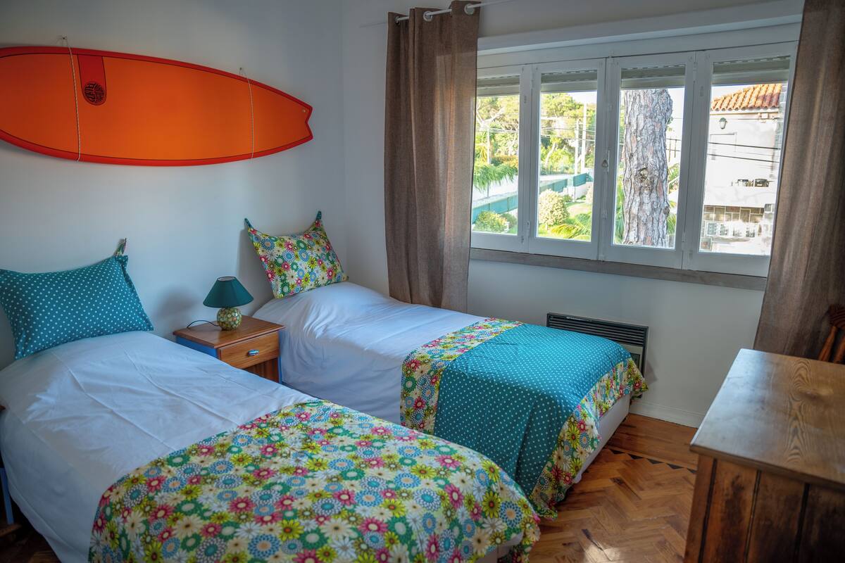 A room with two beds, a surfboard on the wall, a window with curtains, and a wooden table.