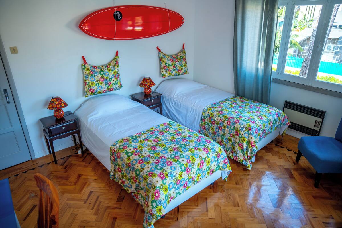 A room with two beds, a red surfboard on the wall, and a window with a view of palm trees.
