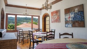 A dining area with a table and chairs, a chandelier, and a view of the countryside through large windows. - Moby Dick Lodge Colares (Sintra)