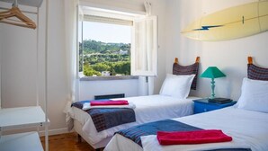 A room with two beds, a window with a view of greenery, a surfboard on the wall, and a small table with a lamp. - Moby Dick Lodge Colares (Sintra)