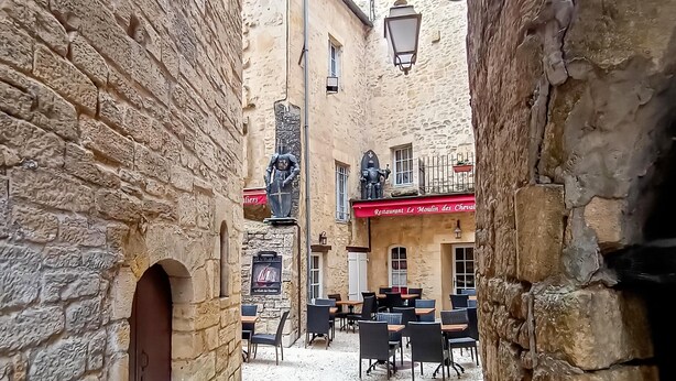 Une ruelle étroite bordée d’immeubles en pierre, avec des tables installées à l’extérieur et une enseigne indiquant la présence d’un restaurant.