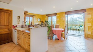 A kitchen with wooden cabinets, a dishwasher, and a dining area with a table and chairs. - Bed and Breakfast 'Chambre Quietude' with Shared Terrace, Shared Garden and Wi-Fi (Saint-Amand de Coly)
