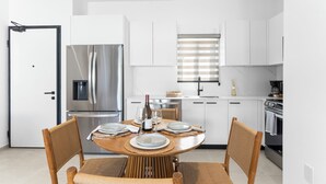 A modern kitchen with white cabinets, a stainless steel refrigerator, a dining table with chairs, and a window with blinds. - Casa Bisani in Cupecoy (Sint Maarten)