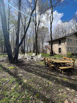 Une table et des chaises en bois pour pique-nique, installées dans une zone boisée, avec une maison en arrière-plan.