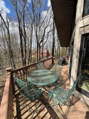 Une terrasse en bois avec une table et des chaises en métal, donnant sur une zone boisée et un bâtiment avec un toit rouge.