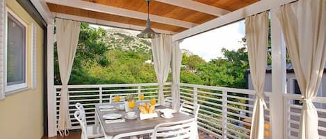 Un espace extérieur abrité, aménagé pour prendre les repas, avec un plafond en bois, des chaises blanches et une table dressée pour le petit-déjeuner.
