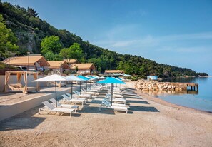 A beach with sun loungers and umbrellas, a rocky shoreline, and a forested hill in the background. - Lanterna Home for 4+2 (Tar)