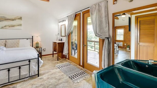 A bedroom with a bed, a nightstand, a desk, a window with curtains, and a bathroom with a bathtub. - Hilltop Lookout #2 at Busch Mt- Stunning Views! (Eureka Springs)