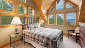 Un dormitorio con cama, una silla, cómoda y amplias ventanas que dan a vistas de árboles y montañas.