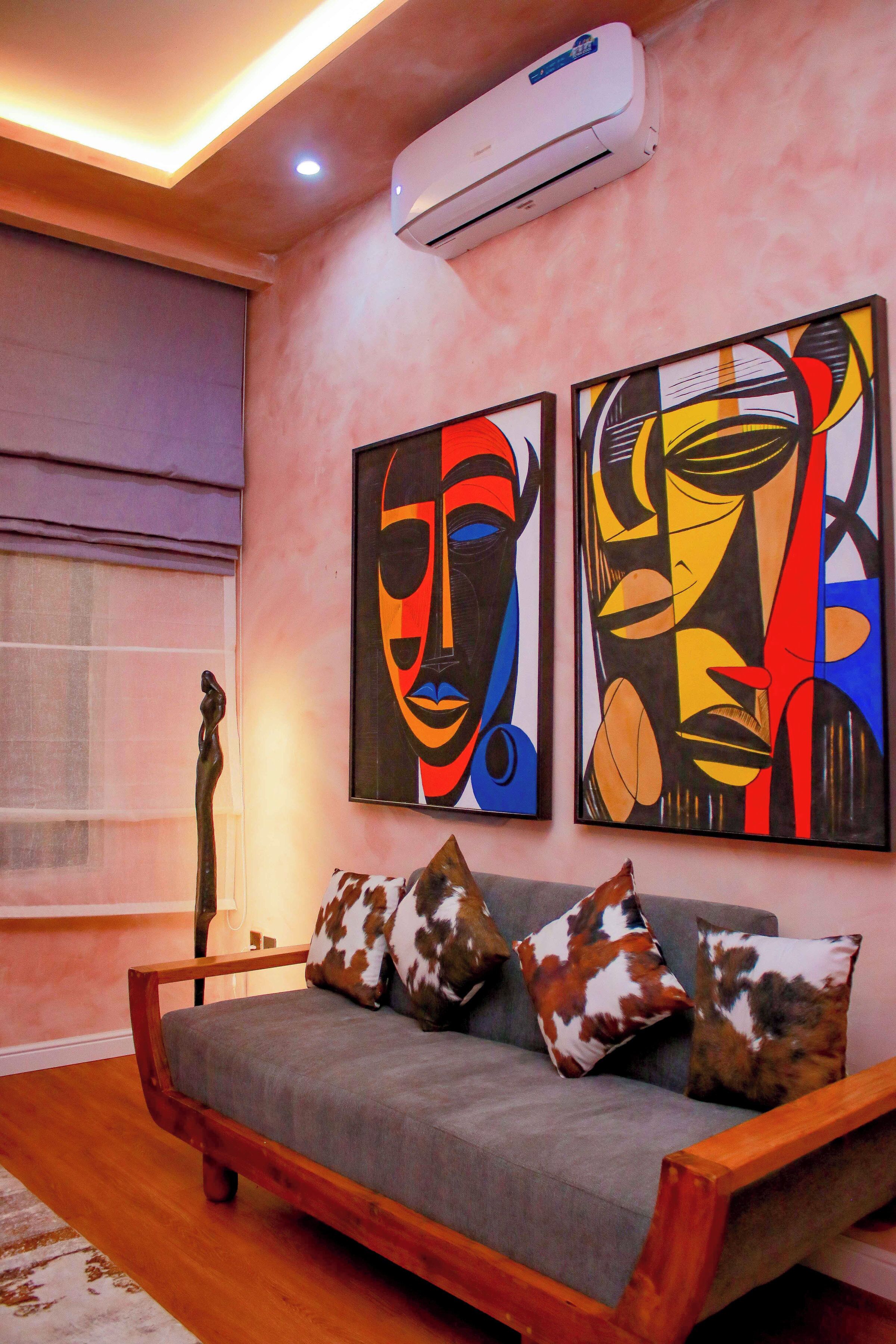 A living room with a grey sofa, wooden frame, and patterned cushions. There are two abstract paintings on the wall, and an air conditioning unit above the sofa.