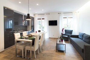A modern kitchen with white cabinets, a dining table with chairs, a flat-screen TV mounted on the wall, and a gray sofa in the living area. - Boba's House, Rome - Apartment St. Peter (Roma)