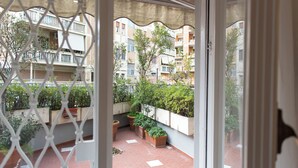 Visão através de uma janela de um pátio com plantas em vasos e piso de tijolos.