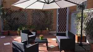 Um pátio externo com móveis de vime, um grande guarda-sol e plantas em vasos.