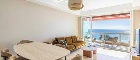 A modern living room with a large round table, comfortable seating, a sofa, and a balcony with a view of the sea.