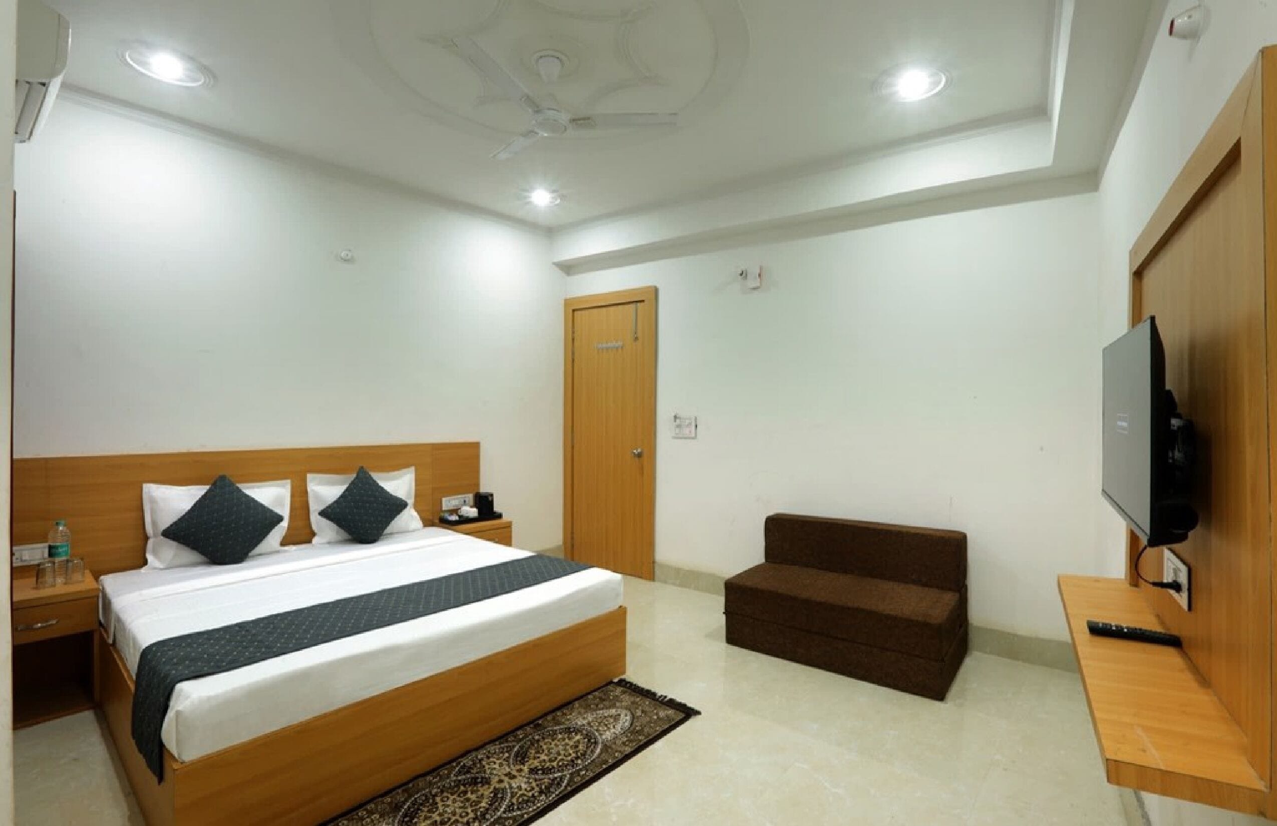A hotel room with a large bed, a wooden headboard, a television, a small sofa, and a ceiling fan.