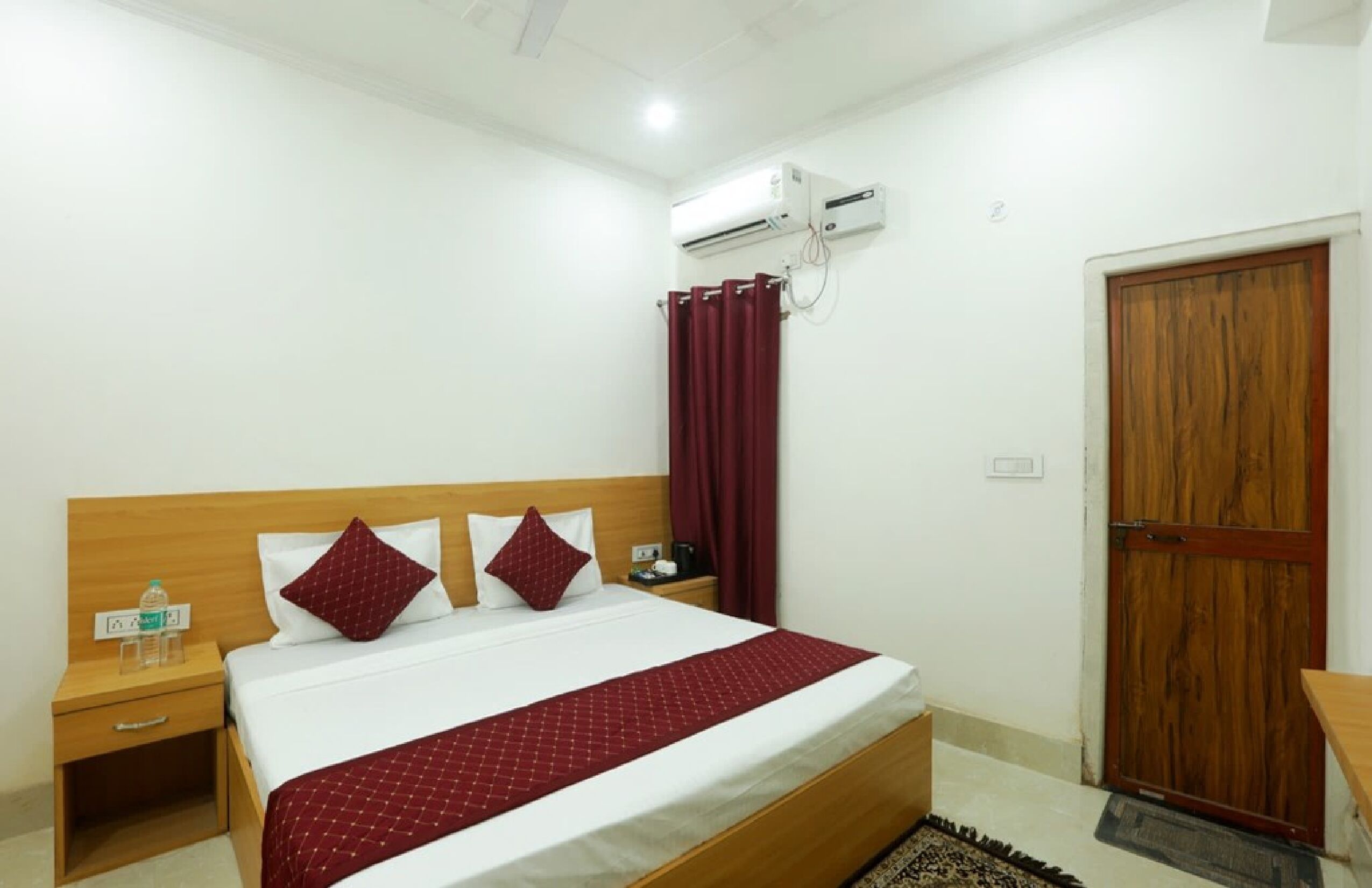 A hotel room with a wooden bed, white bedding, a red throw blanket, a nightstand with a water bottle, an air conditioning unit, and a wooden door.