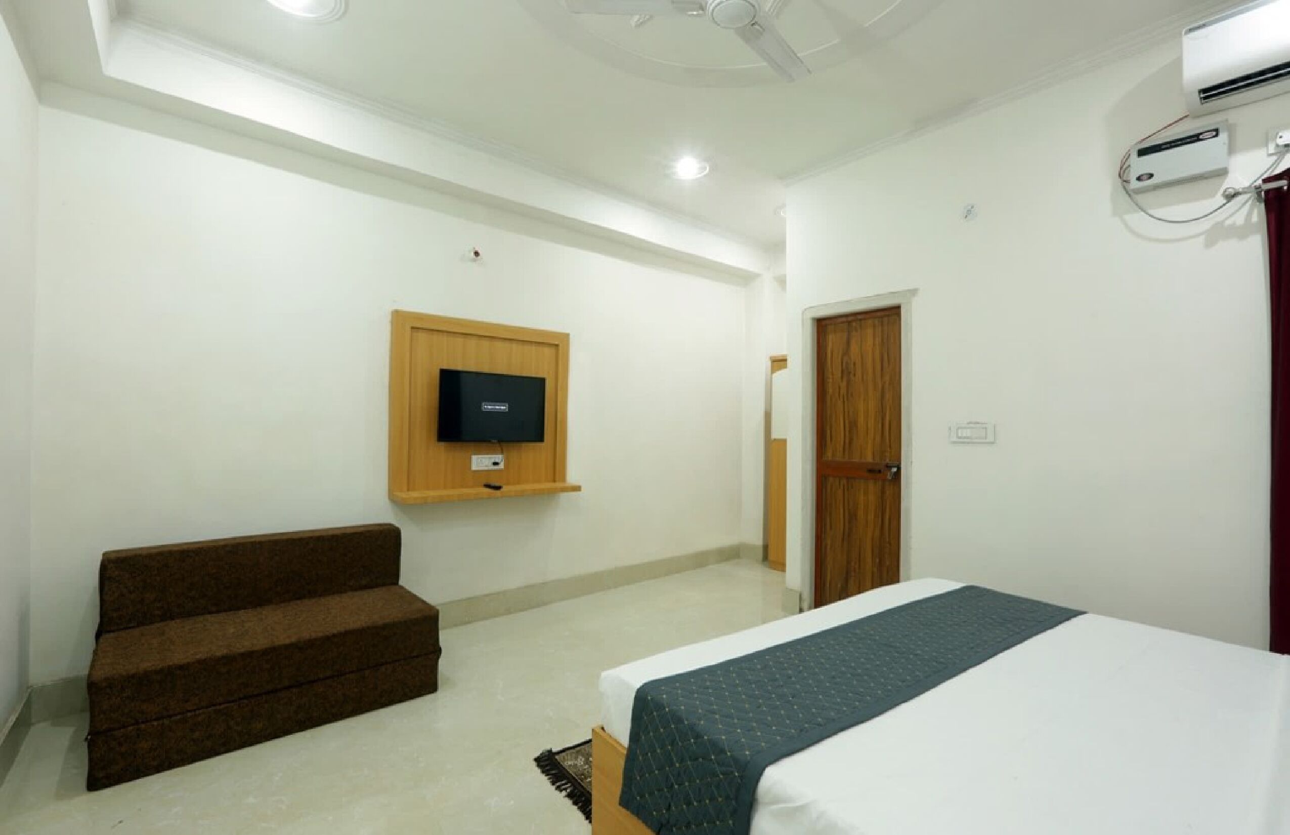 A hotel room with a bed, a TV mounted on the wall, a wooden door, and a ceiling fan.