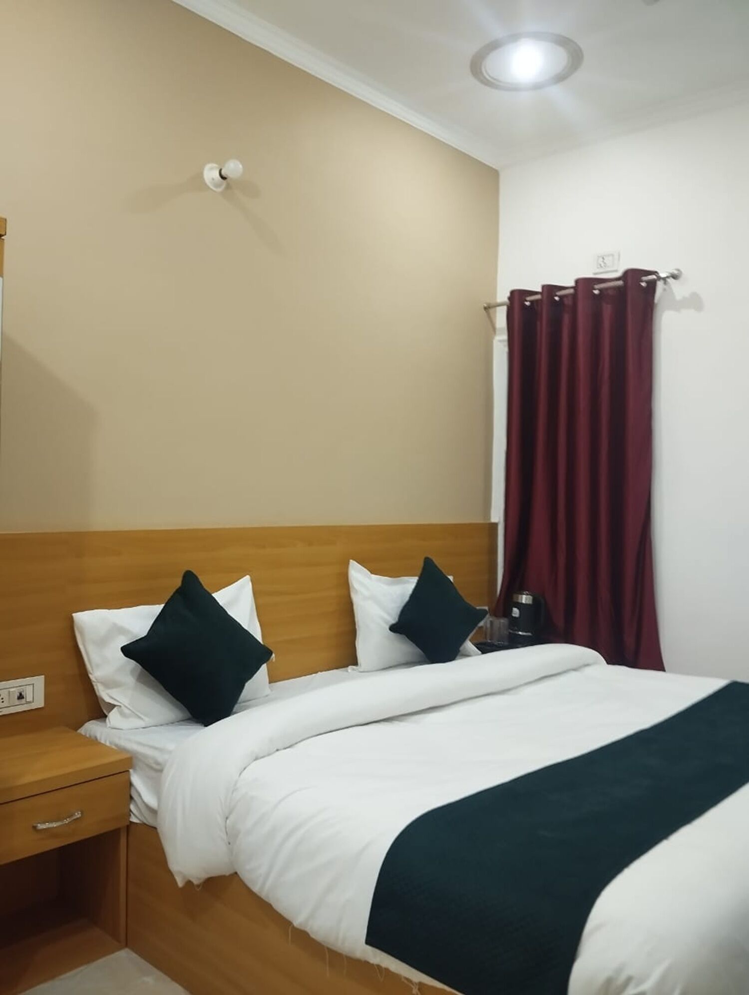 A hotel room with a wooden headboard, two white pillows, a dark green throw blanket, a nightstand with a lamp, and a window with red curtains.