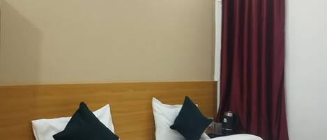 A hotel room with a wooden headboard, two white pillows, a dark green throw blanket, a nightstand with a lamp, and a window with red curtains.