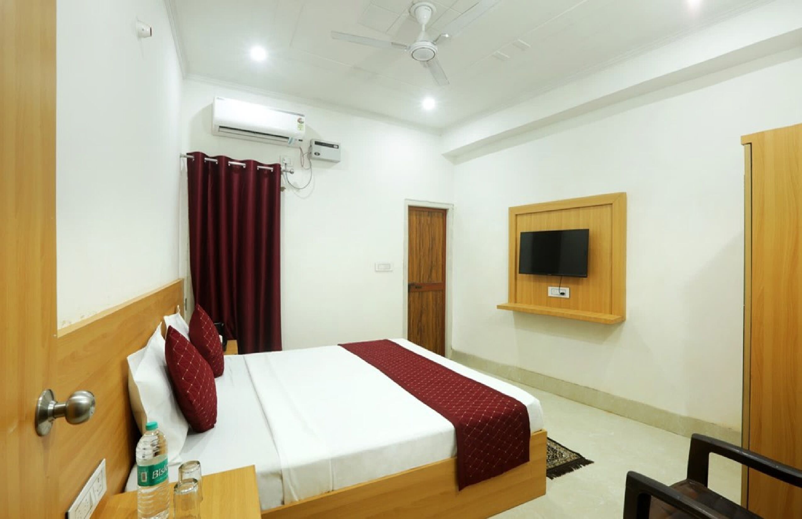 A hotel room with a bed, a TV mounted on the wall, an air conditioning unit, and a wooden door.