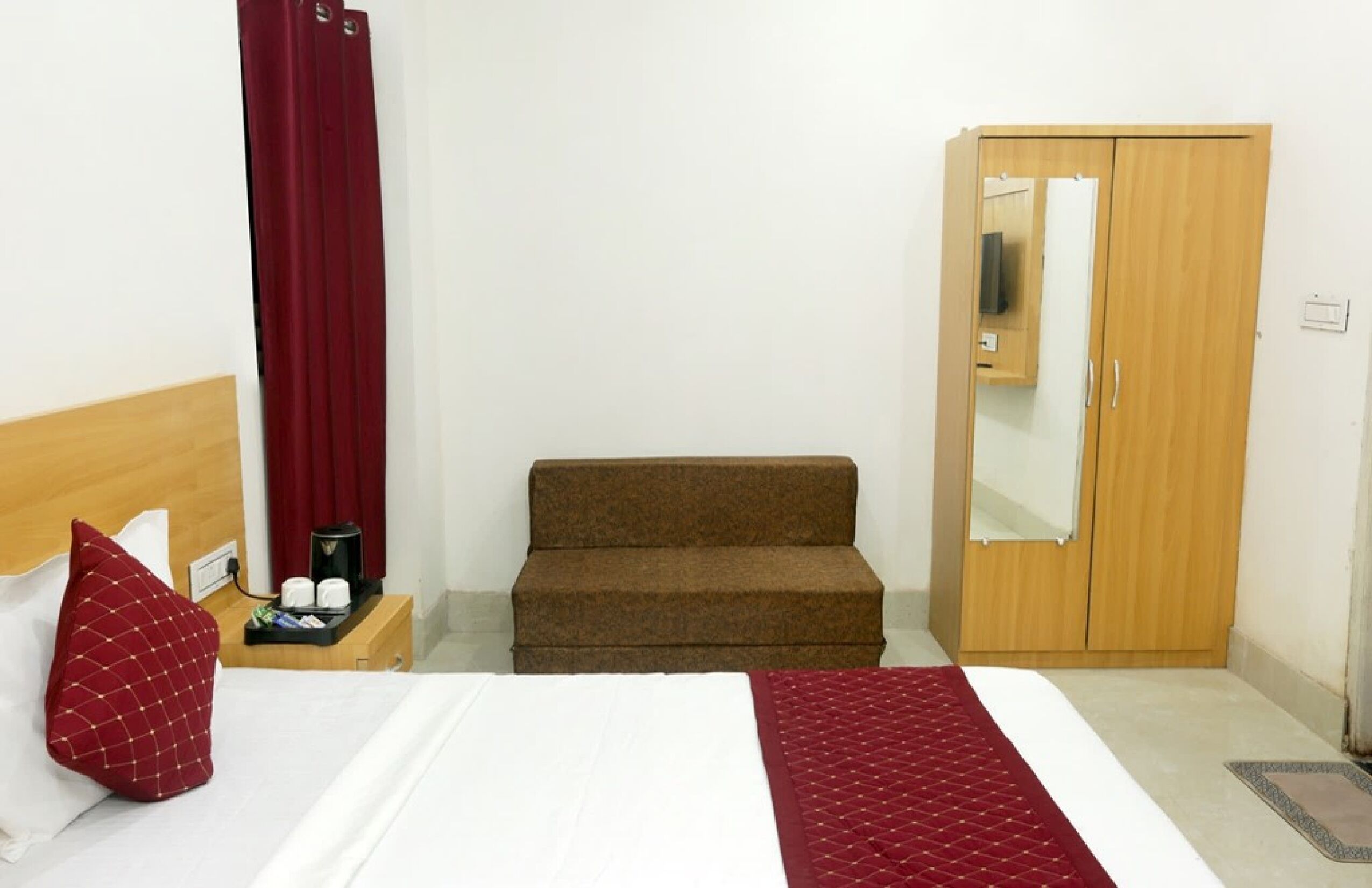 A hotel room with a bed, a wooden wardrobe, a brown sofa, a red curtain, and a small table with a tray.