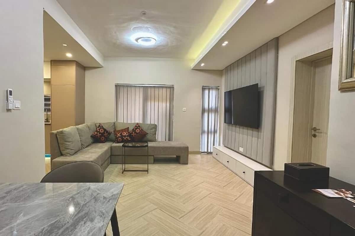 A modern living room with a sectional sofa, a small table, and a flat-screen TV mounted on the wall.