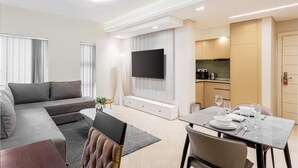 A modern living room with a grey sofa, a flat-screen TV mounted on the wall, a dining table set with plates and glasses, and a kitchen area with cabinets and appliances. - DOMO by Design Union (Lekki)