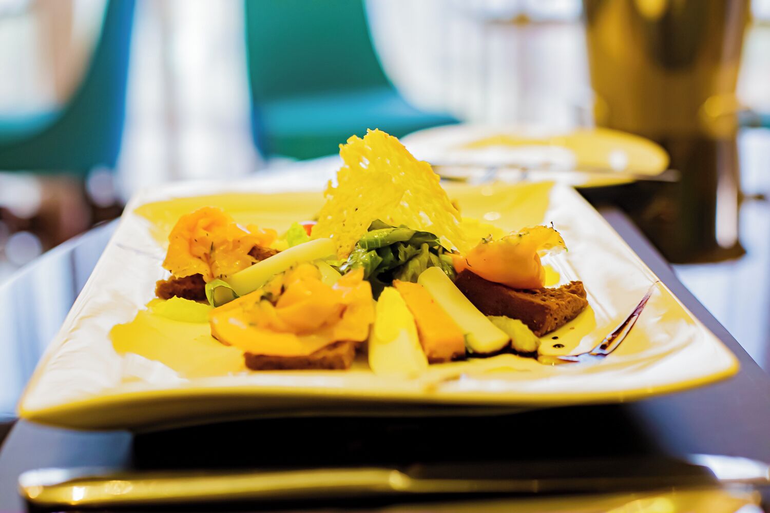 A square plate with a dish consisting of a slice of bread topped with a yellow sauce, green vegetables, and a piece of meat.